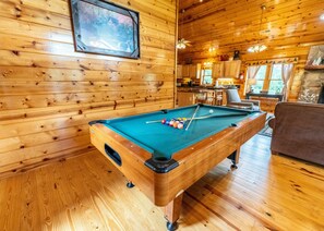 A pool table in a wooden cabin with a kitchen and seating area visible in the background. - Little Sugar Camp (Sevierville)
