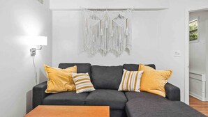 A modern living room with a grey sofa, yellow and striped cushions, a wooden coffee table, and a macrame wall hanging. - The Hoggtown Hideaway (Gainesville)