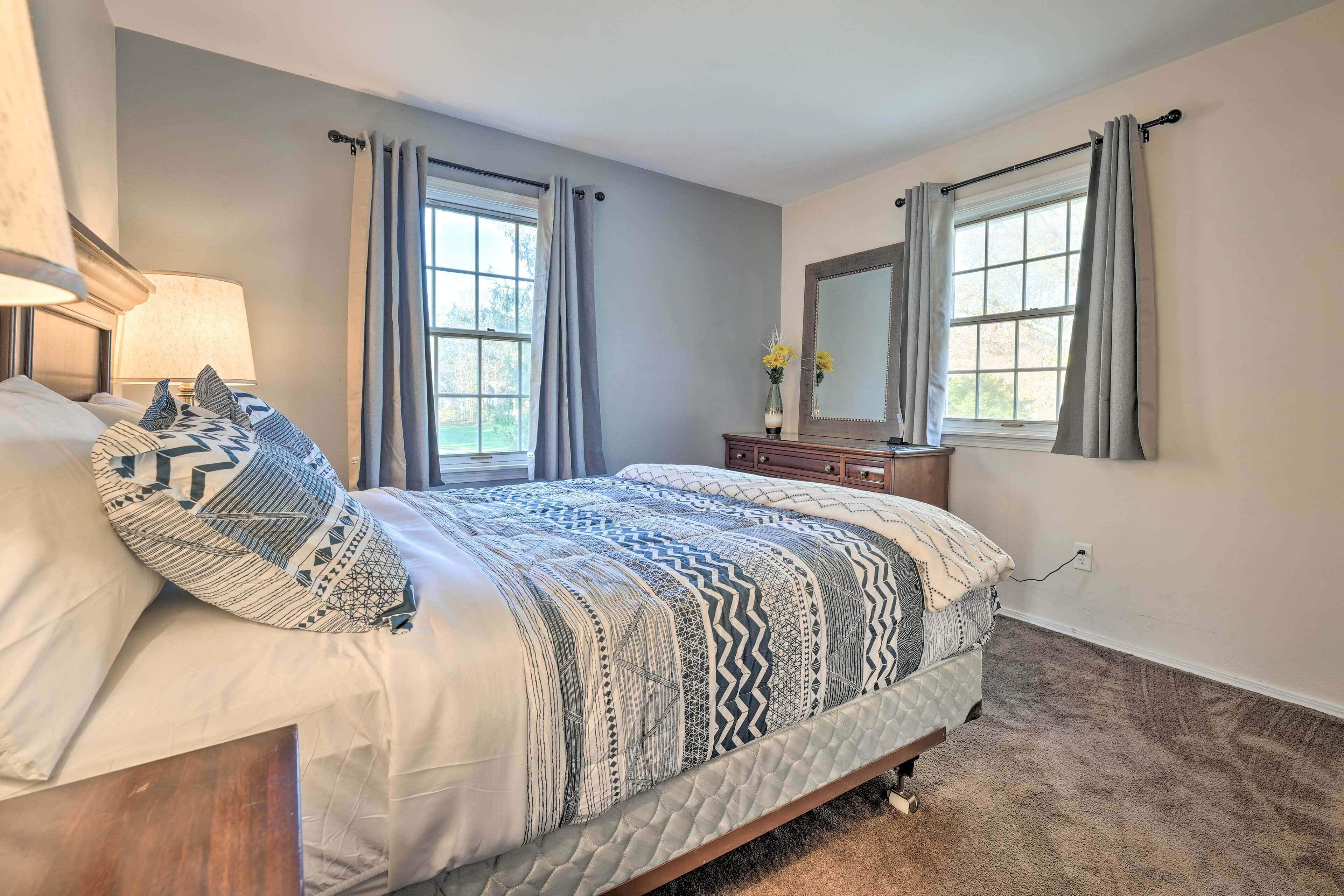 A bedroom with a large bed, two windows with curtains, a dresser, a mirror, and a lamp.