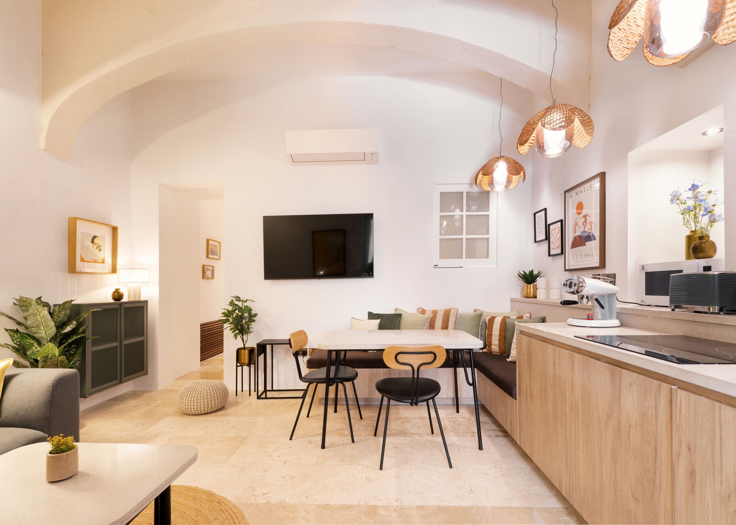 A modern kitchen with a dining area, a TV mounted on the wall, and a large window.