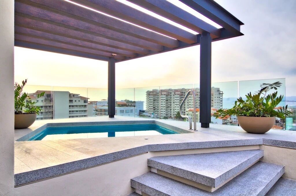 A rooftop pool area with a view of high-rise buildings, a pergola, and potted plants.