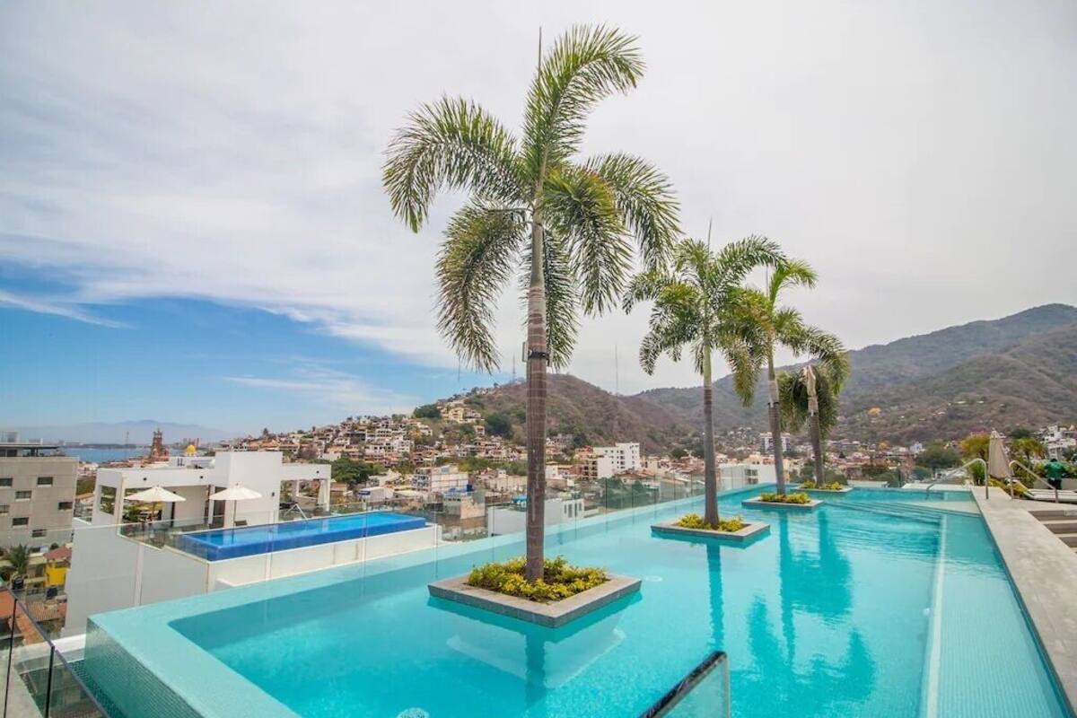 A rooftop infinity pool with palm trees, overlooking a coastal town and mountains.