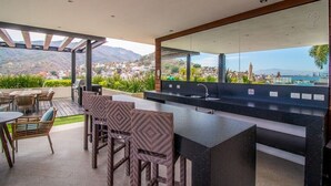 A modern outdoor kitchen with a bar area, seating, and a view of a mountainous landscape. - Condominios Z (Puerto Vallarta)