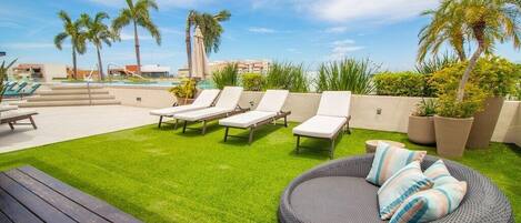 A rooftop lounge with white lounge chairs, a round wicker sofa, and a pool in the background.