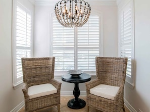 Une pièce d’angle avec deux fauteuils en osier et une table, un lustre et des volets blancs.