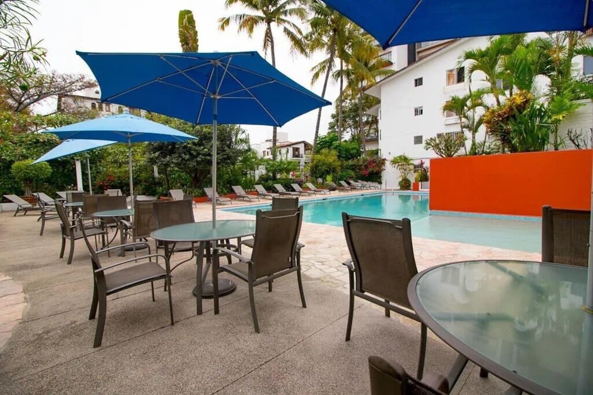 Zona de piscina con sillones de descanso, sombrillas y una mesa de vidrio.