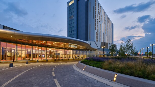 Entrada moderna de hotel com marquise curva, um edifício alto ao fundo e área ajardinada com caminhos.