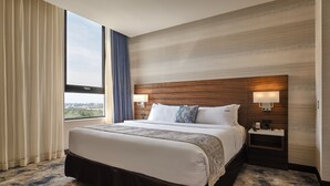 Quarto de hotel com uma cama grande, duas mesas de cabeceira, um criado-mudo, uma janela com cortinas e vista para a cidade.