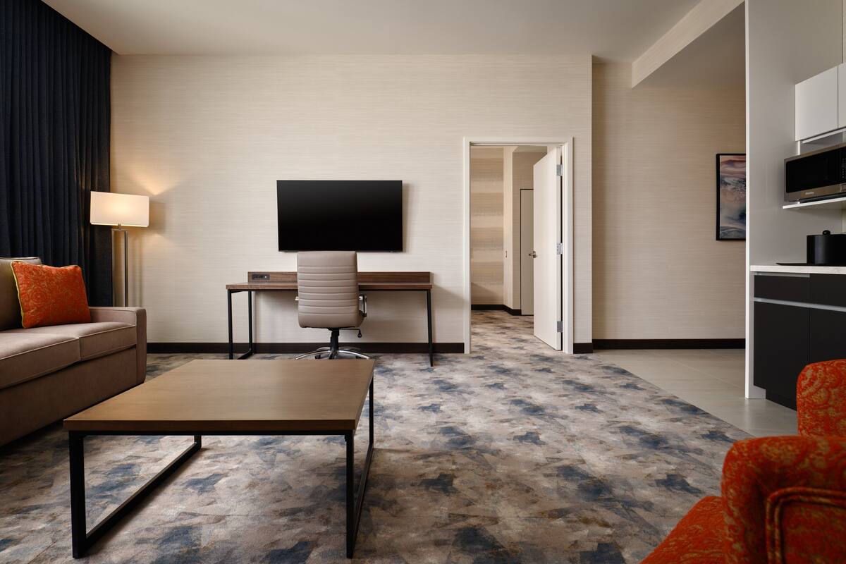 Una habitación de hotel moderna con sofá, una mesa de centro, escritorio, televisor de pantalla plana y kitchenette.