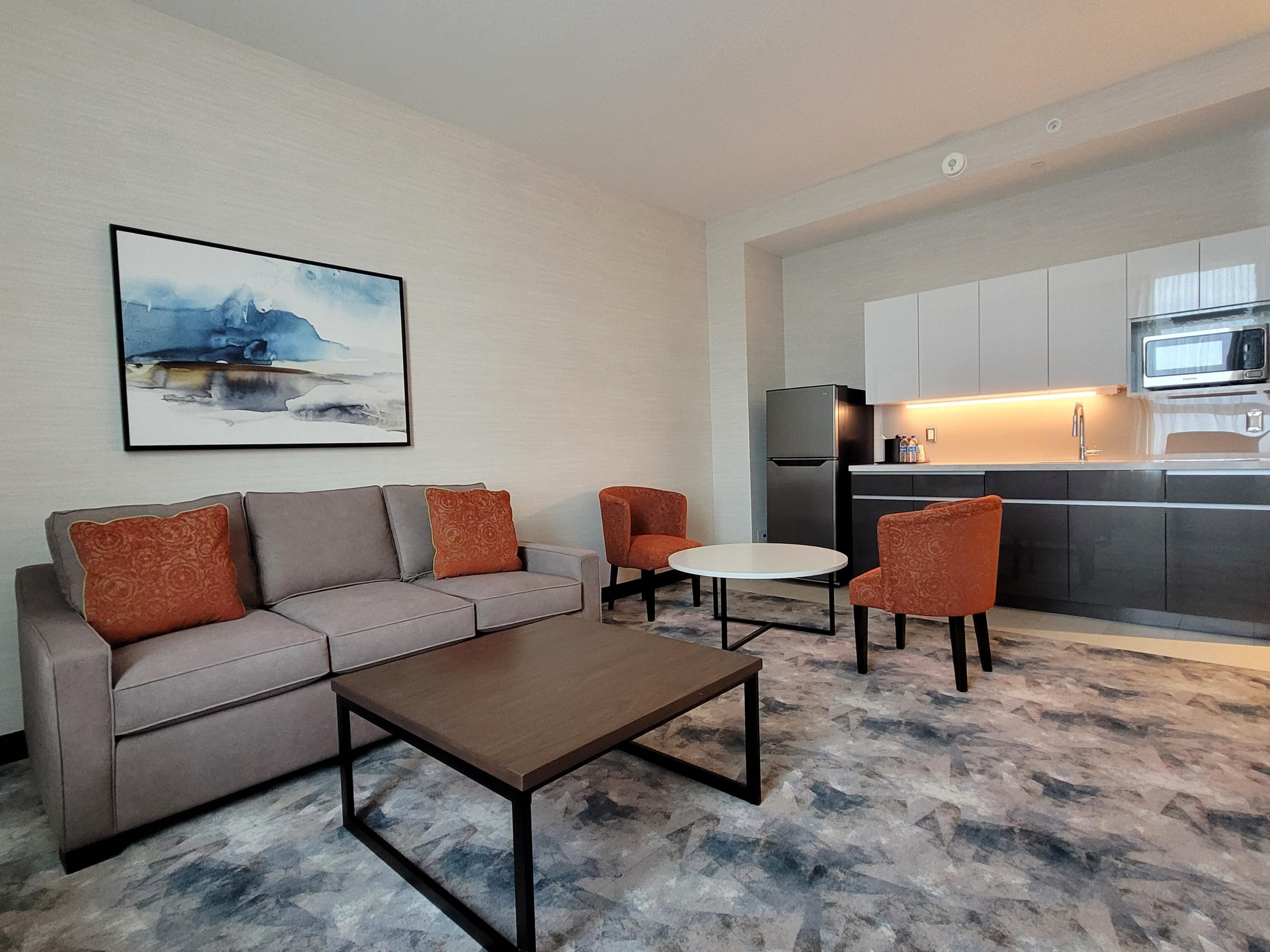 Una sala de estar moderna con un sofá, una mesa de centro y dos sillones. Se ve una cocina con un refrigerador y un microondas al fondo.