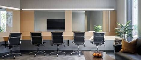 A conference room with a large screen, four black chairs, a coffee table, and a sofa with a plant.