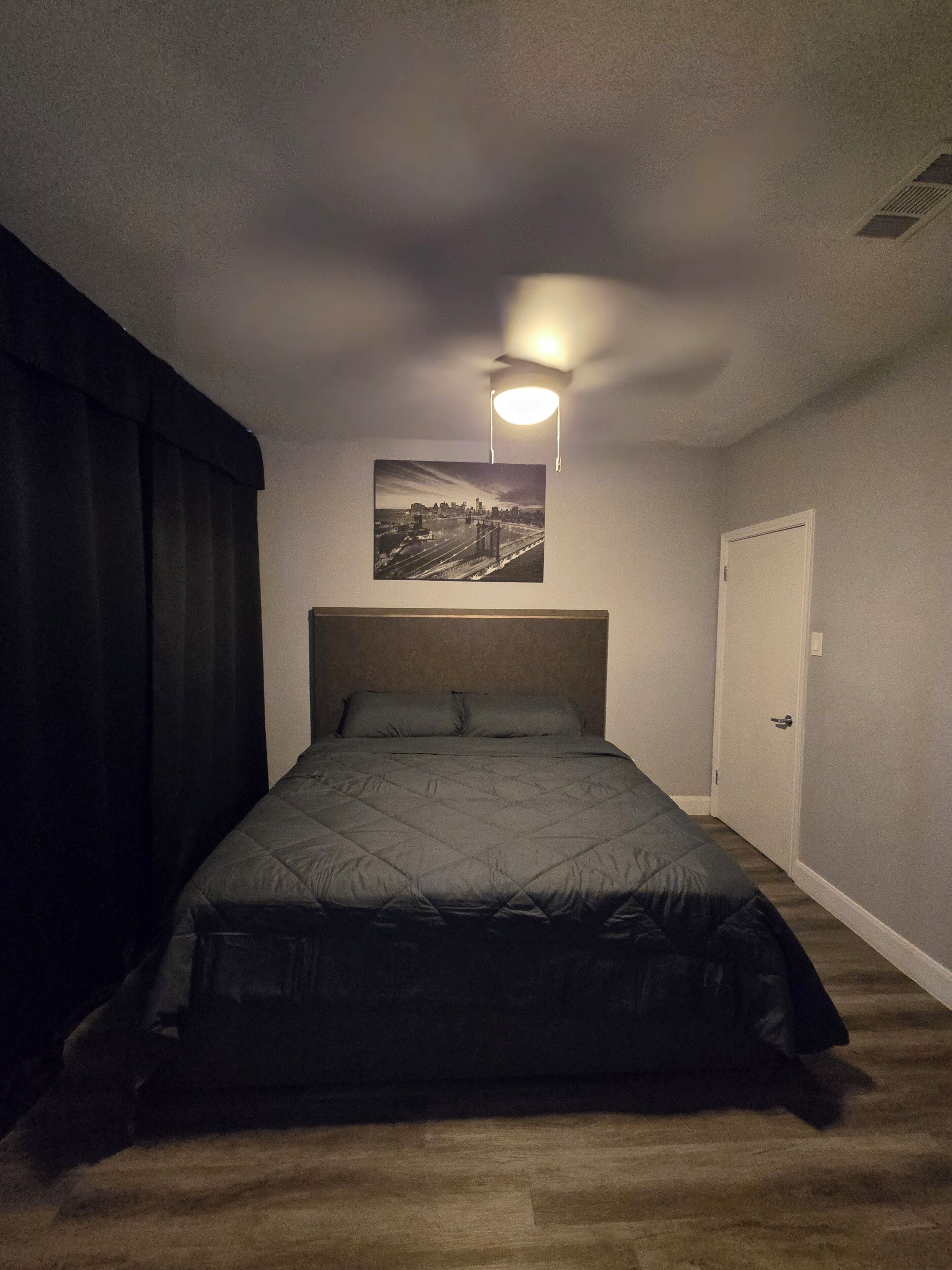 Un dormitorio con cama, una lámpara de techo, una puerta y un cuadro con un paisaje urbano en la pared.