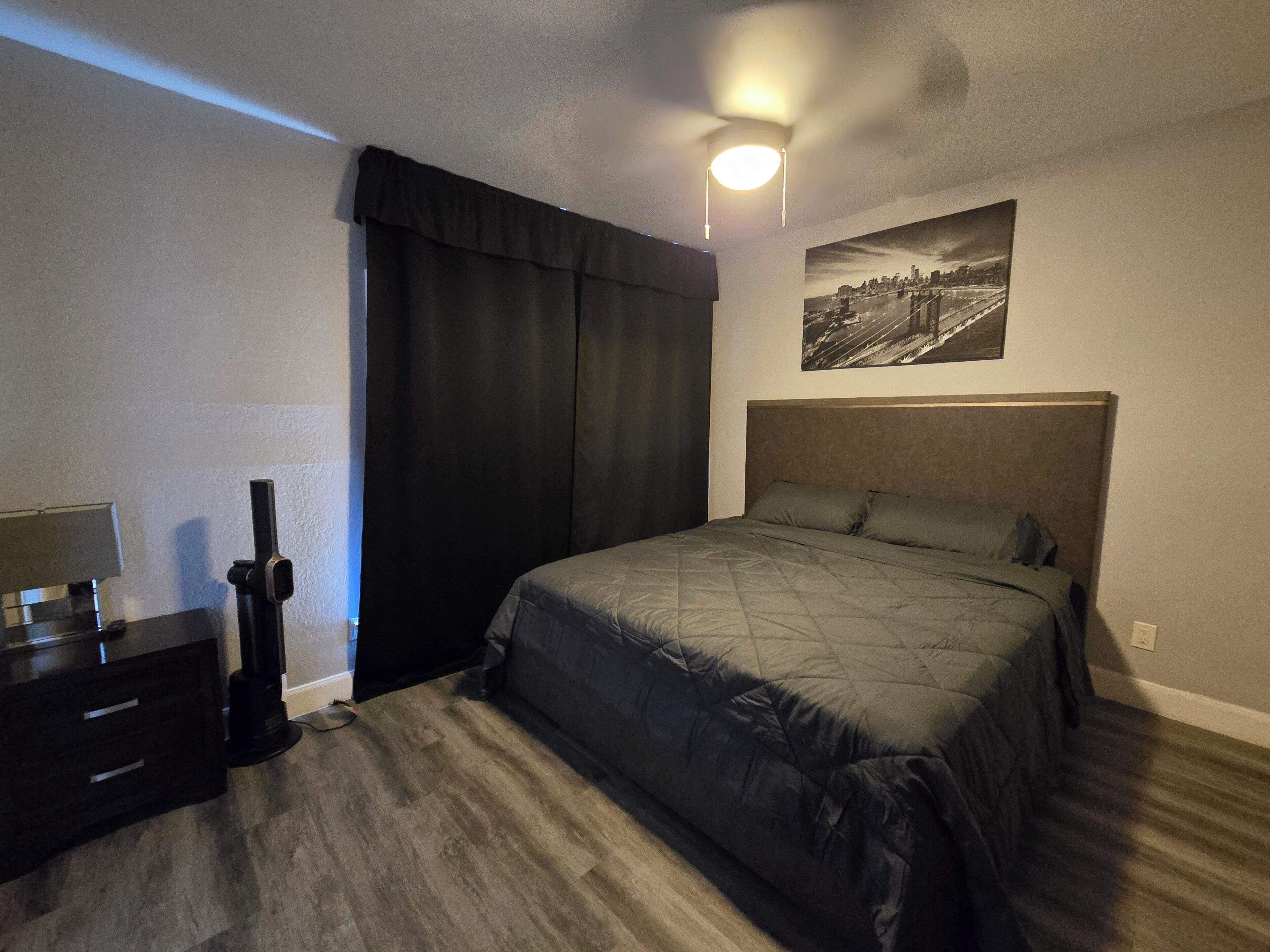Une chambre à coucher comprenant un lit, une table de chevet, une lampe, un luminaire au plafond, une fenêtre avec des rideaux et un tableau accroché au mur.