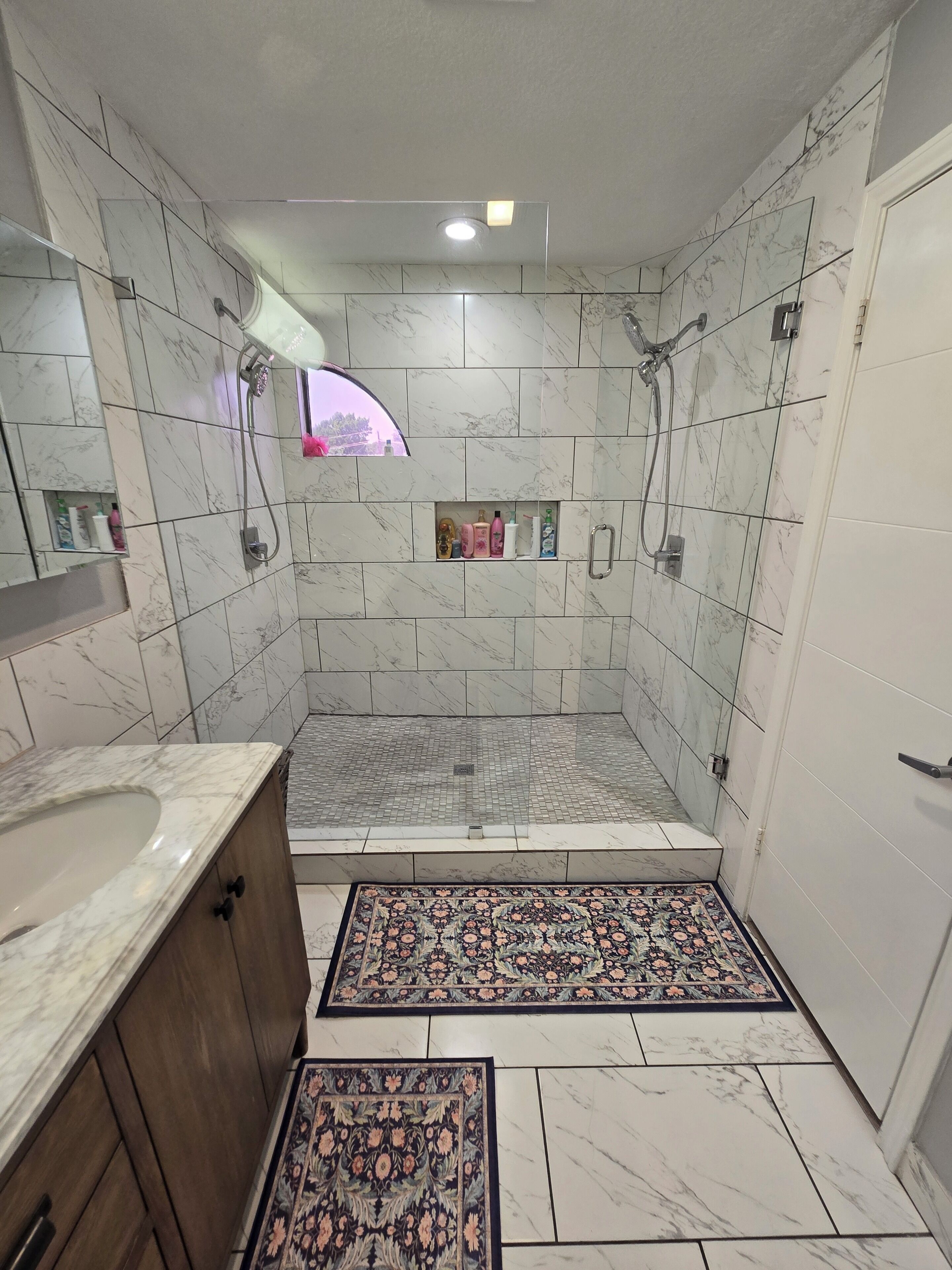 A bathroom with a shower, a sink, a mirror, and a small shelf with toiletries.