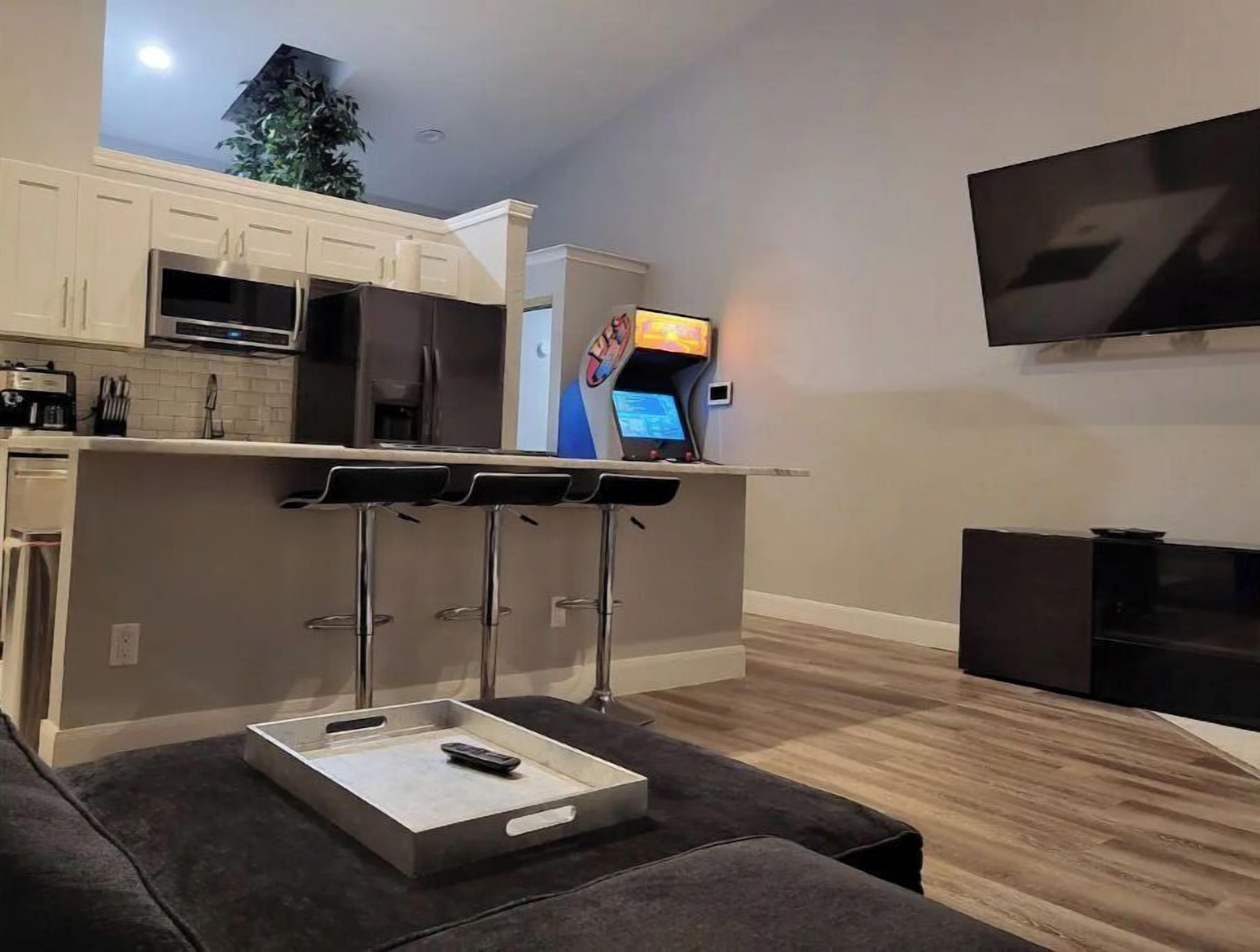 Una cocina moderna con microondas integrado, horno y un refrigerador pequeño. Hay una barra con taburetes y un televisor montado en la pared.