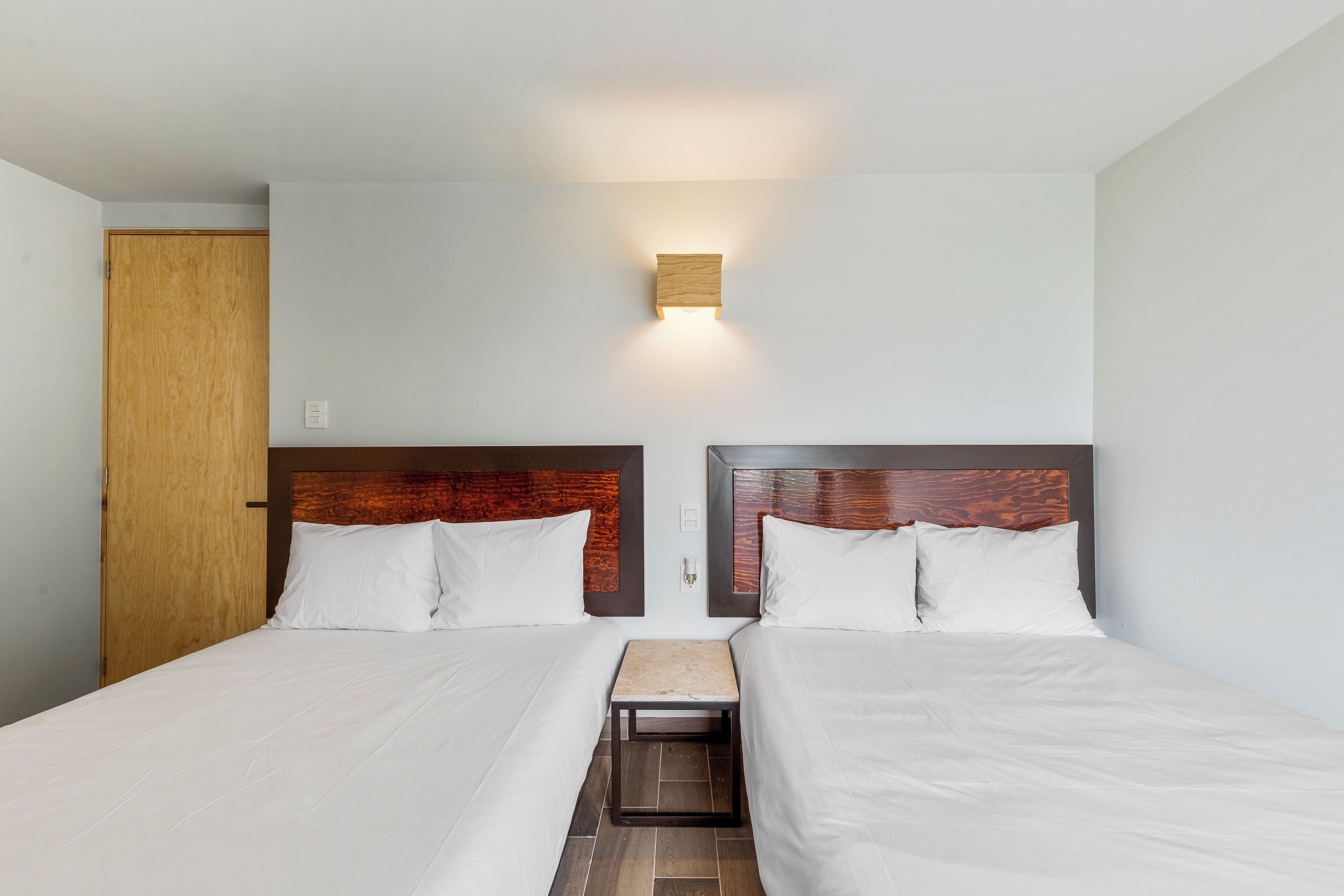 A hotel room with two beds, a wooden door, a small table, and a wall-mounted light fixture.