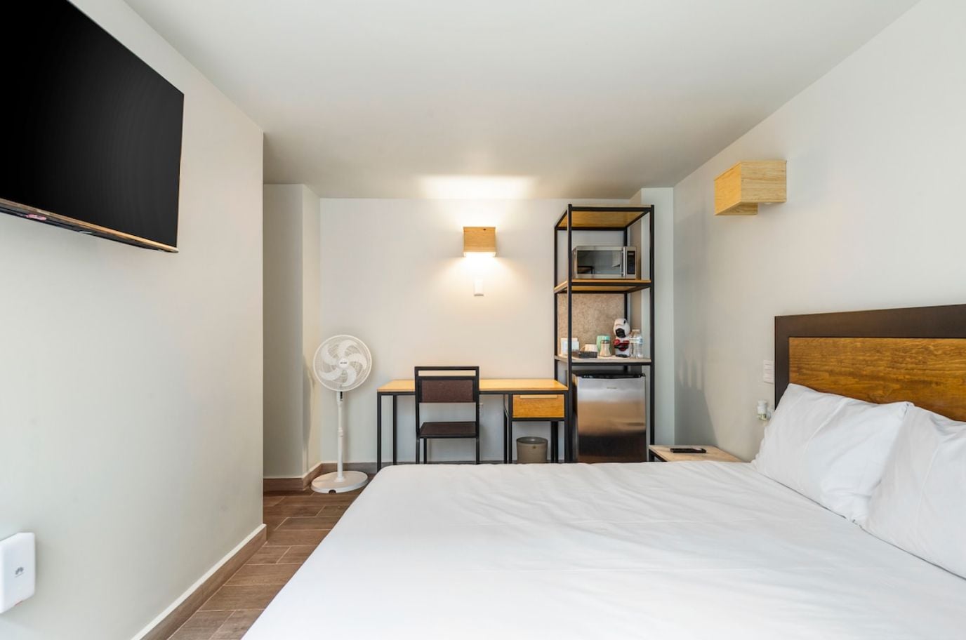 A hotel room with a bed, a desk, a chair, a microwave, a refrigerator, a fan, and a TV mounted on the wall.