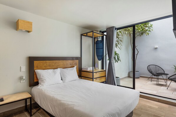 Un dormitorio moderno con cabecera de madera, cama con ropa de cama blanca, una mesita de noche pequeña y vistas a un espacio exterior con sillas y una planta.