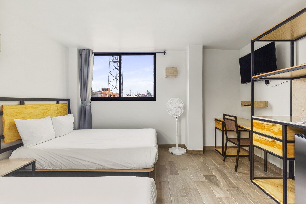 Habitación de hotel con dos camas, un escritorio, una silla, un televisor y una ventana con vista a edificios.