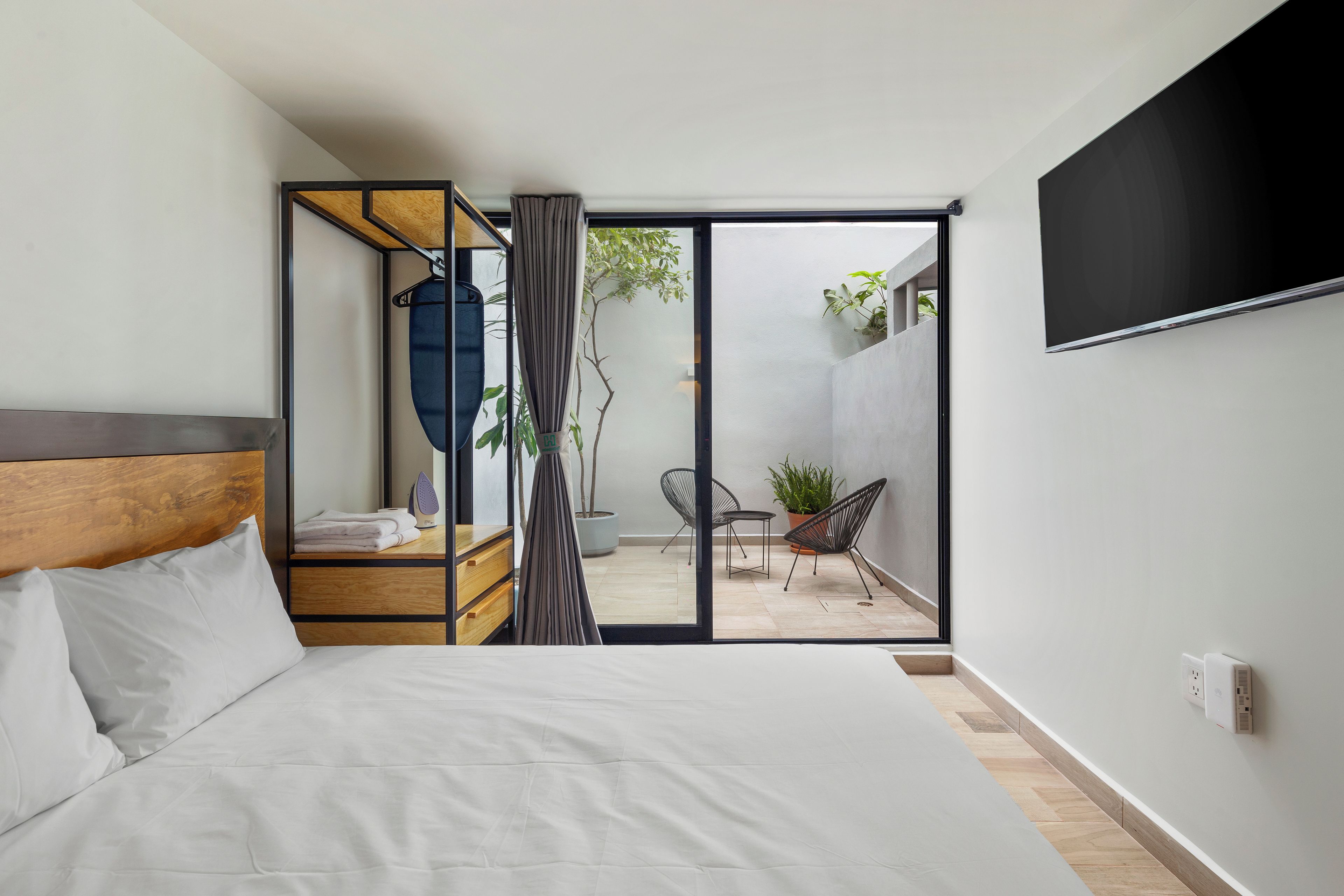 A modern hotel room with a bed, a wooden headboard, a TV mounted on the wall, a balcony with chairs and a potted plant, and a sliding door leading to an outdoor area.