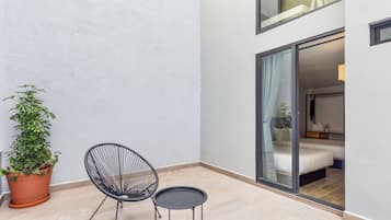 A small outdoor seating area with a chair and table, a potted plant, and a view into a bedroom with a bed and a nightstand.
