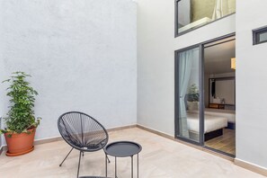 Una pequeña zona de estar al aire libre con una silla y una mesa, una planta en maceta y vistas a un dormitorio con cama y mesita de noche.