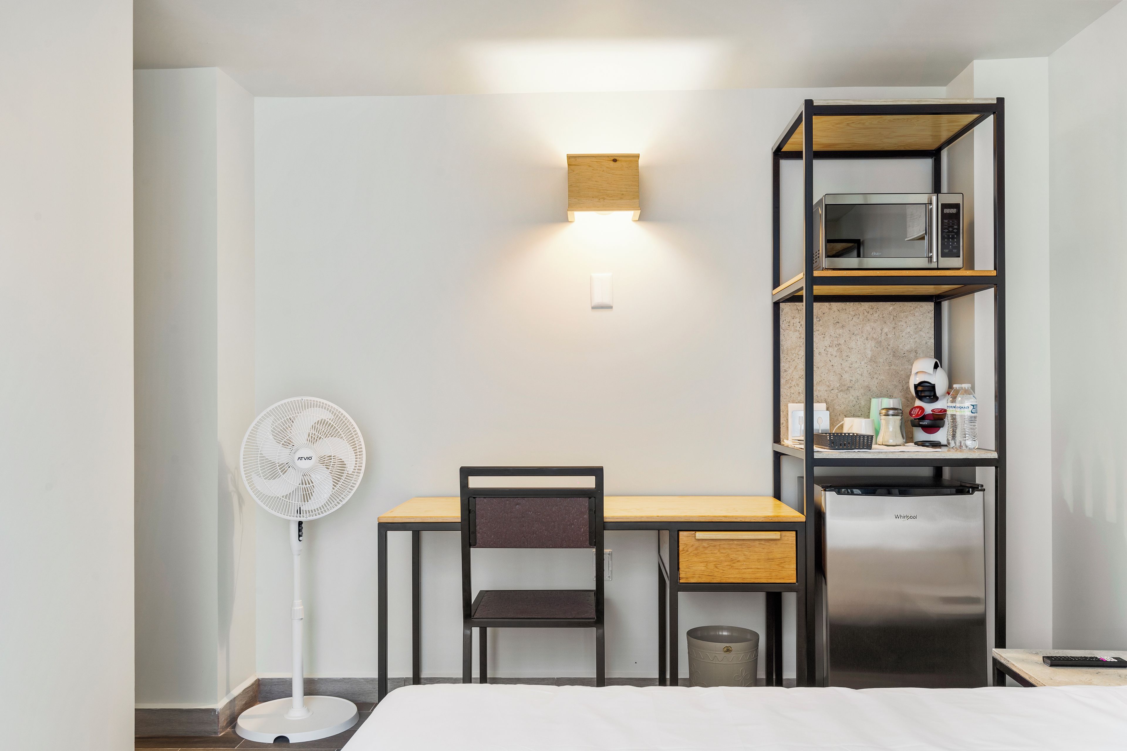 Ein kleines Hotelzimmer mit einem Bett, einem Schreibtisch mit Stuhl, einem Standventilator, einer Mikrowelle, einem Kühlschrank und einer kleinen Mülltonne.