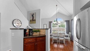 Una cocina con armarios de madera, un refrigerador de acero inoxidable, un reloj y una zona de comedor con mesa y sillas.