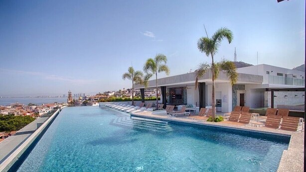 A swimming pool with lounge chairs and palm trees, overlooking a cityscape. - Condominios AR (Puerto Vallarta)