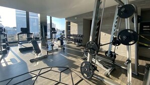 A modern gym with various exercise equipment, large windows offering a city view, and a spacious layout. - Condominios AR (Puerto Vallarta)