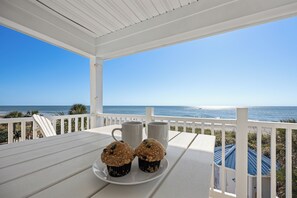 A white wooden deck with a view of the ocean, two white mugs, and two muffins on a white plate.