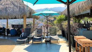 A terrace with thatched umbrellas, lounge chairs, and a view of mountains. - Condominios 180PR (Puerto Vallarta)
