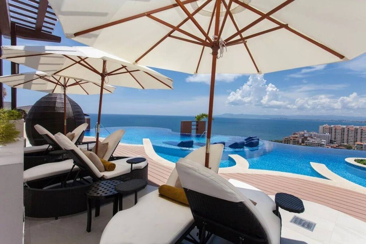 Zona junto a la piscina con sillones de descanso y sombrillas, con vistas a una piscina infinita y al océano.