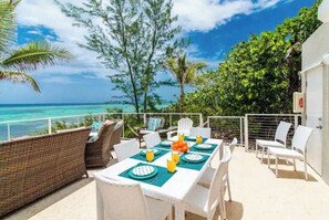 An outdoor dining area with a view of the ocean, white wicker furniture, and a table set with plates and orange juice. - Ocean's Edge: North Side Grand Cayman Beachfront Villa (North Side)