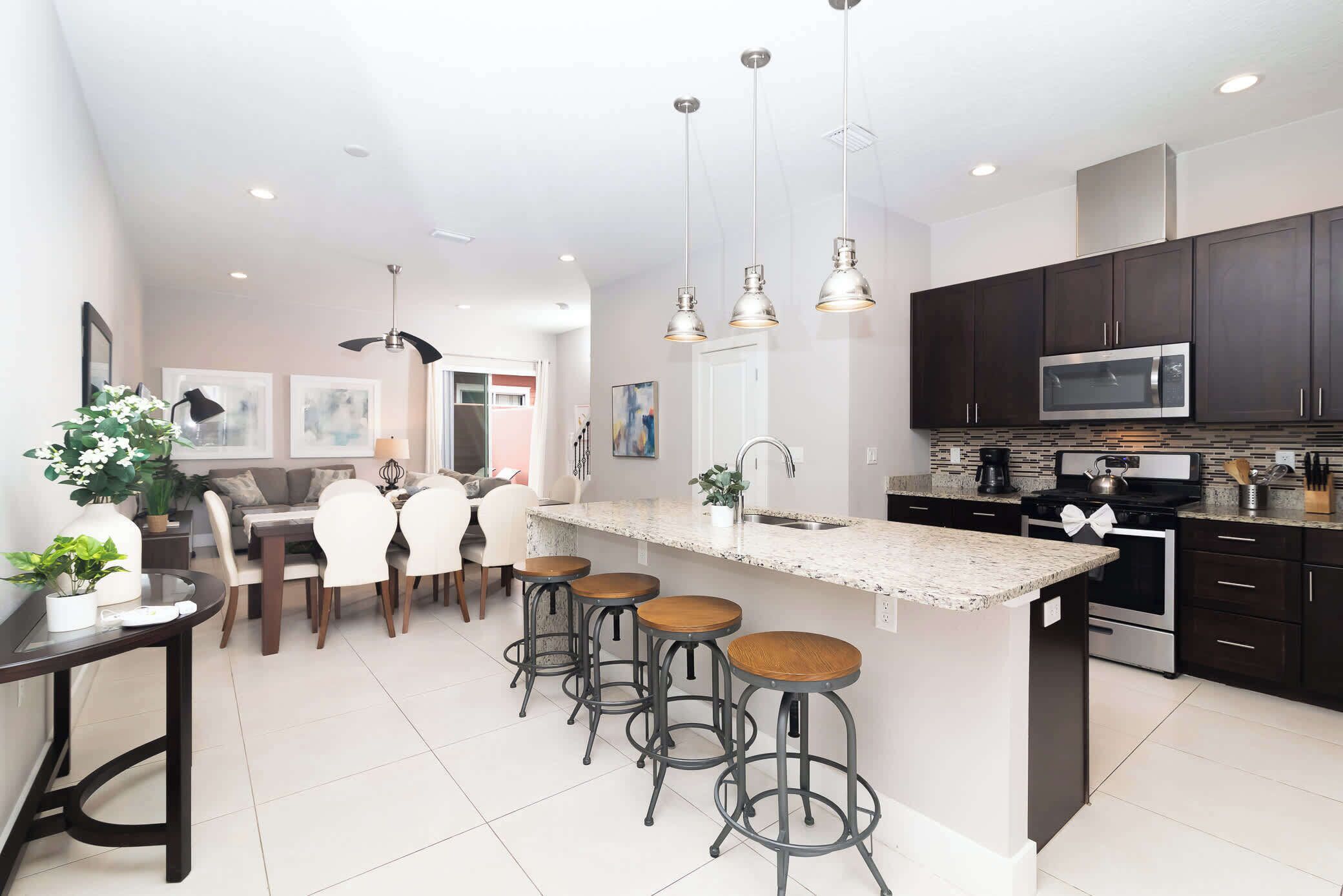 A modern kitchen with dark cabinets, a stainless steel microwave, and a granite countertop. There are four stools at the island and a dining area with a table and chairs.