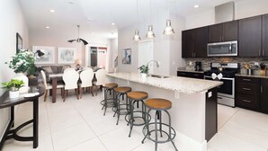Una cocina moderna con armarios oscuros, un microondas de acero inoxidable y una encimera de granito. Hay cuatro taburetes en la isla y un comedor con mesa y sillas.