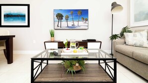 Un salón con una mesa de centro de vidrio, un control remoto, una jarra de agua y un cuenco de fruta. En la pared, hay un cuadro de una playa, una lámpara y un sofá con un cojín.