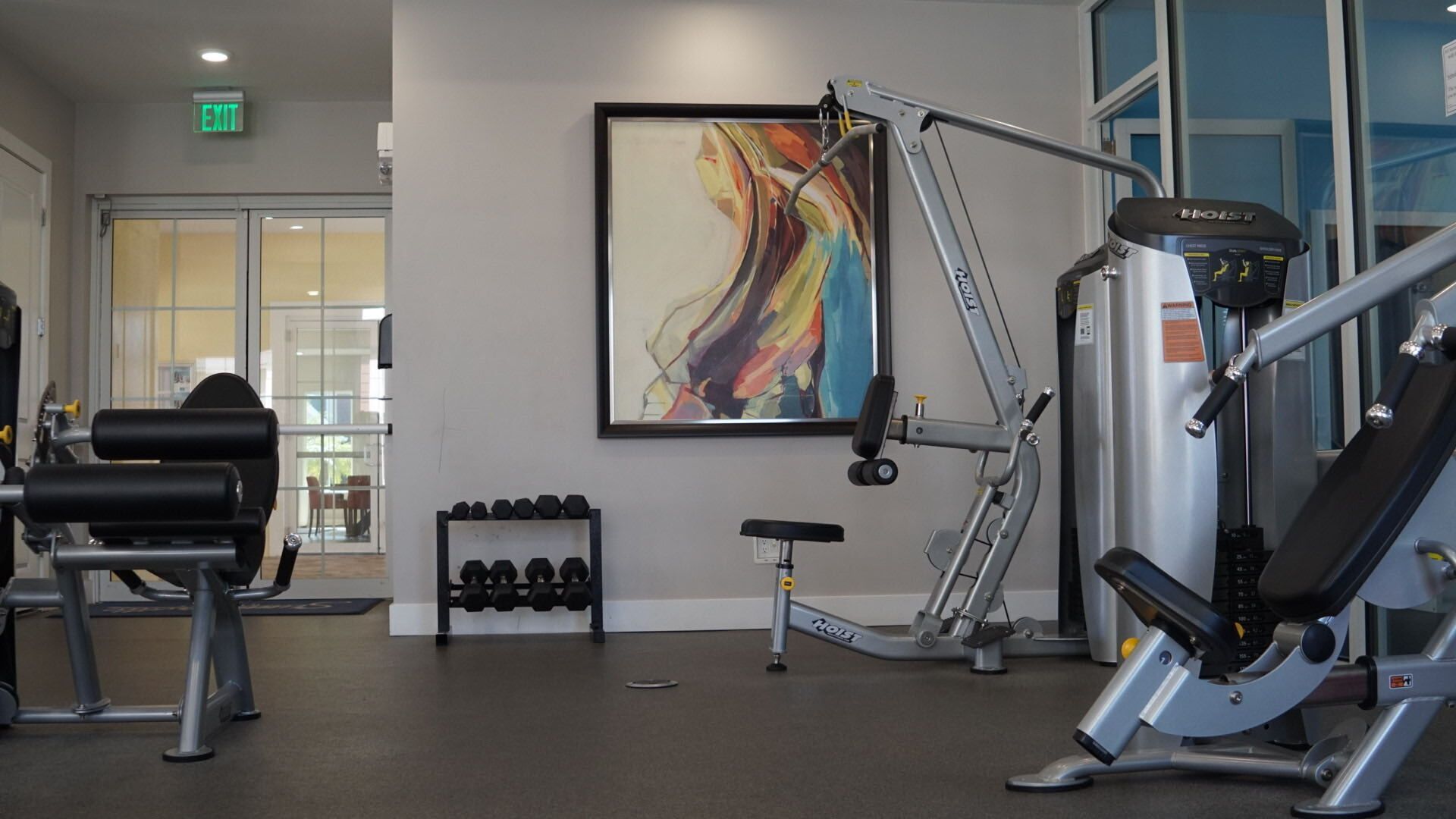 Un gimnasio con diverso equipo de ejercicio, un estante de mancuernas y un cuadro abstracto y colorido en la pared.