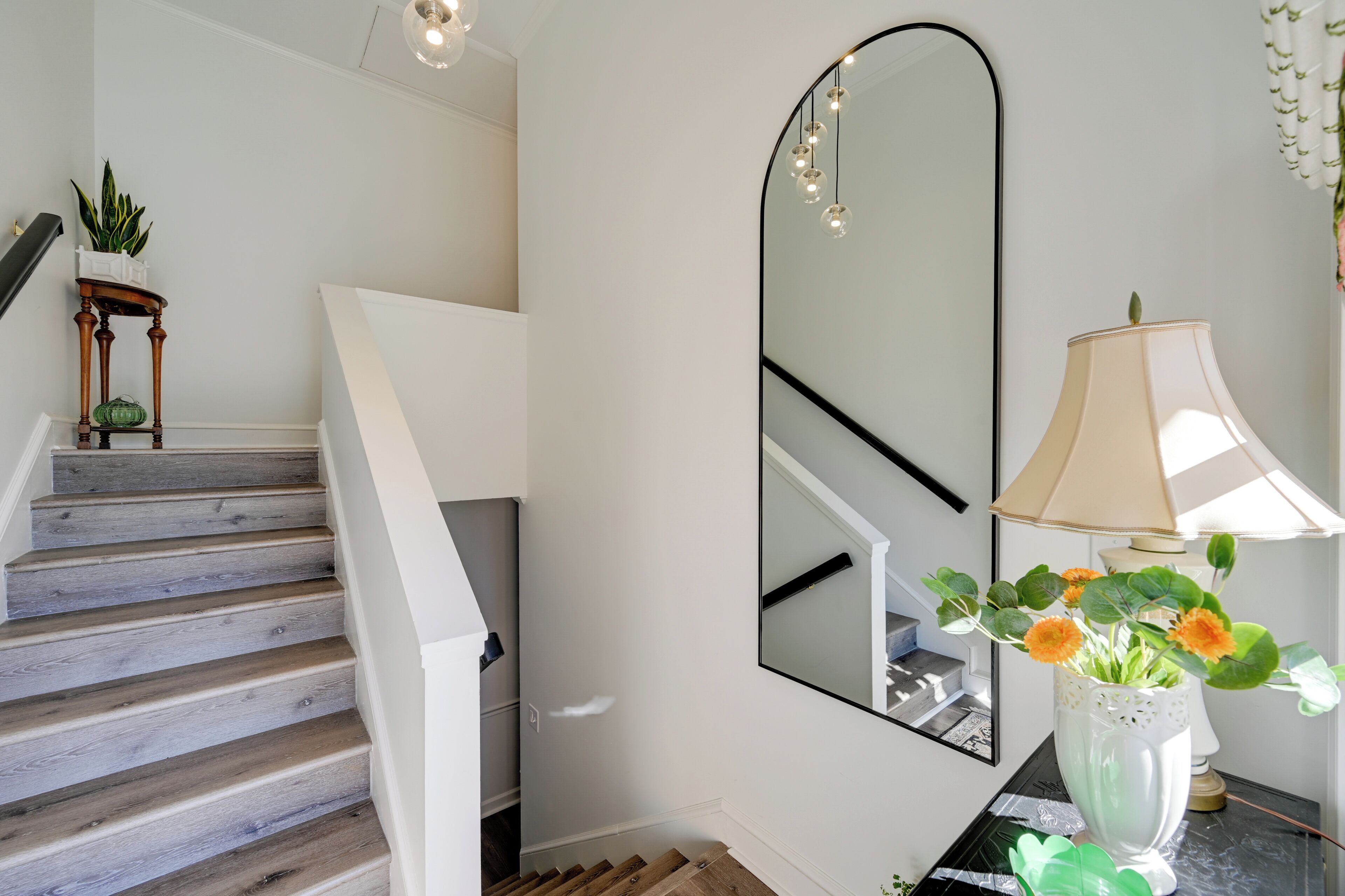 Un escalier avec des marches en bois, un mur blanc, un miroir, une lampe et un vase rempli de fleurs.