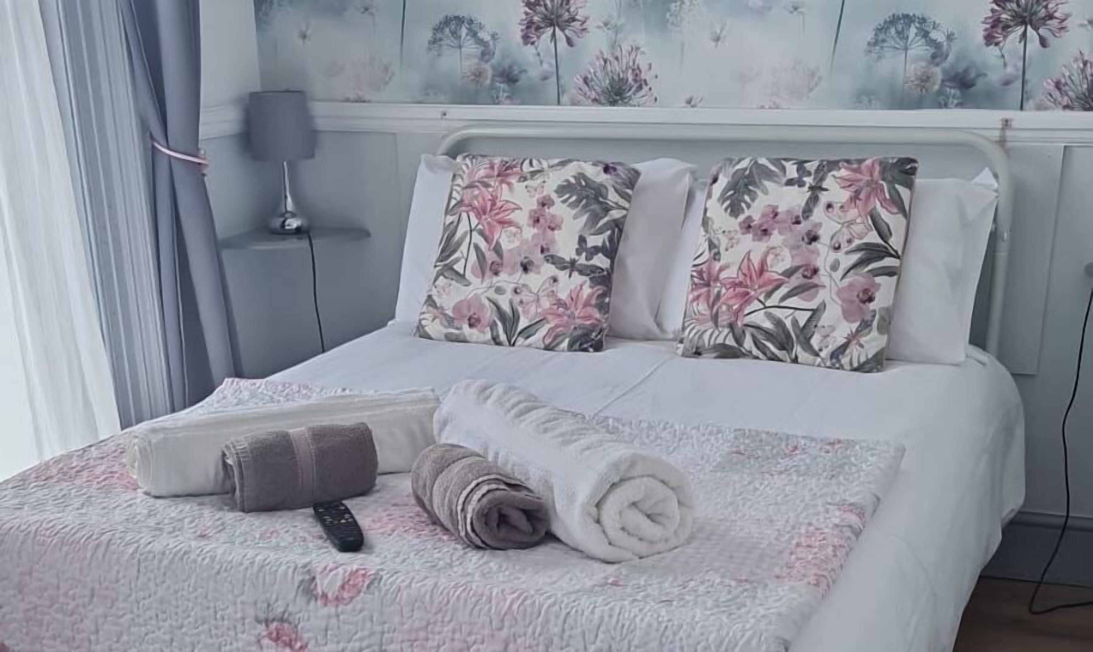 Una cama bien hecha con almohadas de estampado floral, una toalla enrollada y un control remoto sobre una colcha con motivos florales.