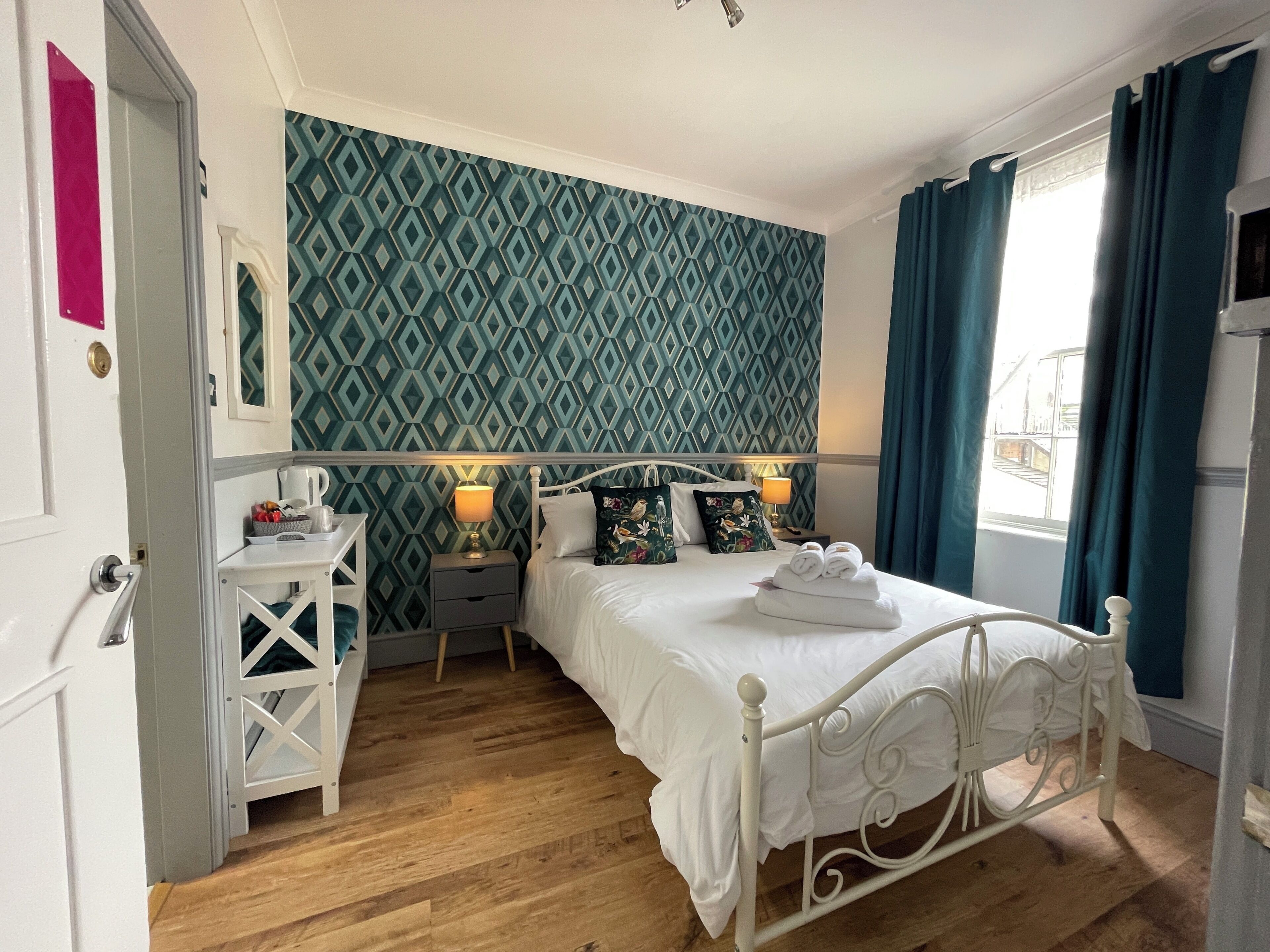 A neatly arranged bedroom with a patterned wallpaper, a white bed, and a window with curtains.