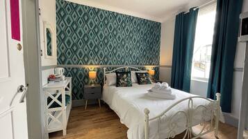 A neatly arranged bedroom with a patterned wallpaper, a white bed, and a window with curtains.