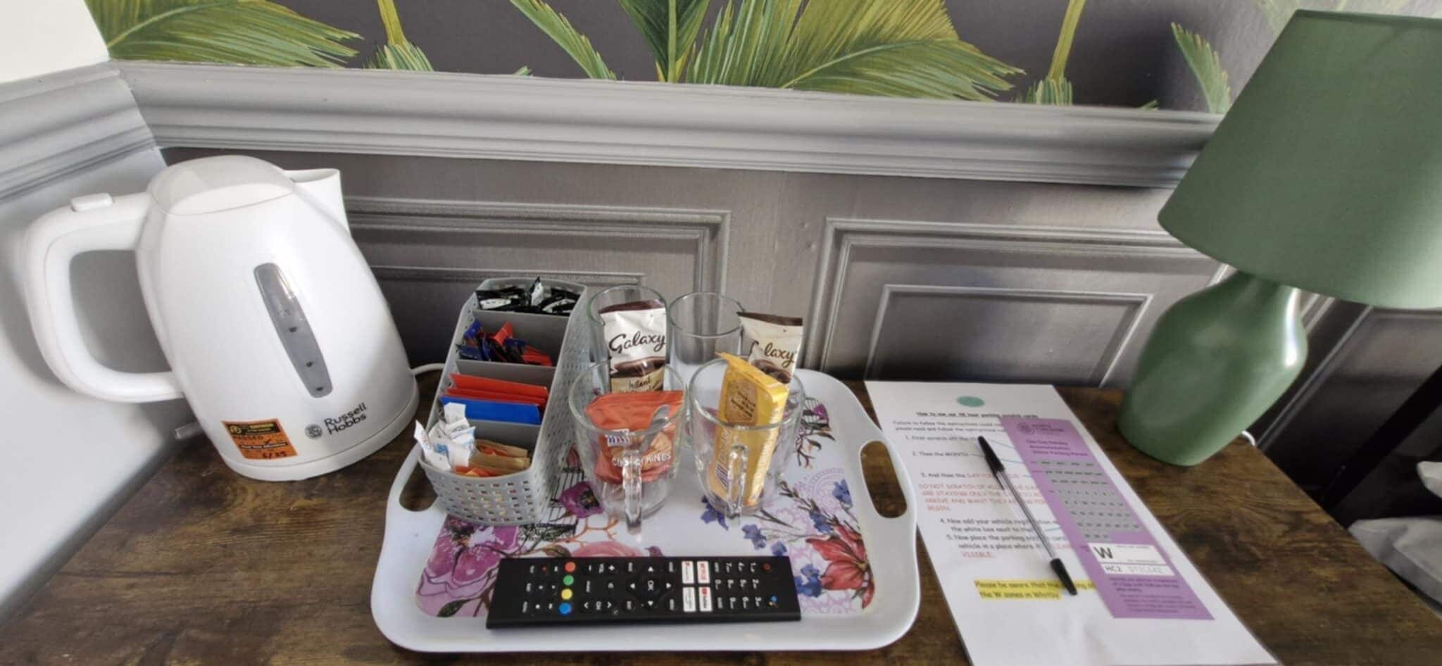 Un escritorio con hervidor, un control remoto, una bandeja con aperitivos y una lámpara.