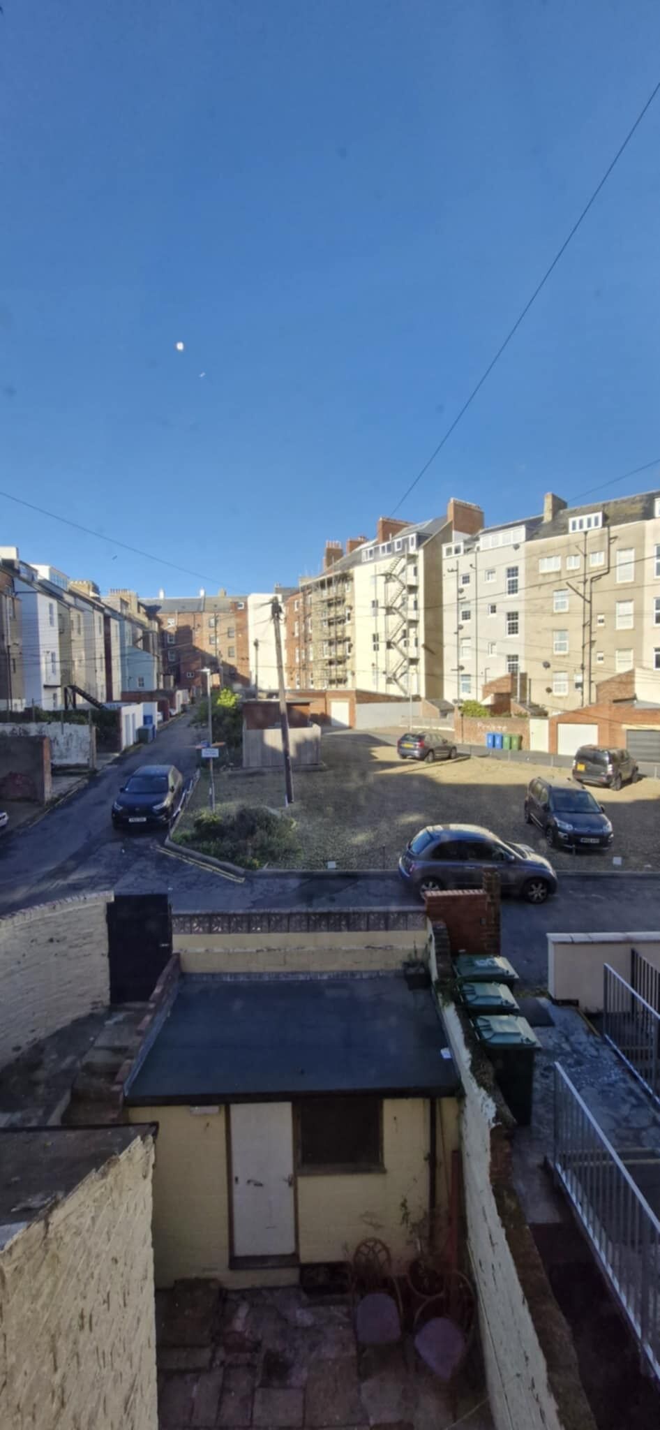 A residential street with multiple parked cars, a small building with a flat roof, and a clear blue sky.