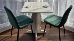 A corner room with a white table and two teal chairs, large windows with sheer curtains, and a view of buildings outside.