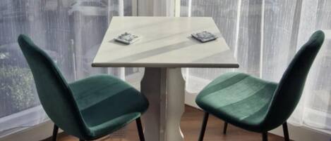 Habitación de esquina con una mesa blanca y dos sillas azul turquesa, ventanas amplias con cortinas traslúcidas y vista a edificios.