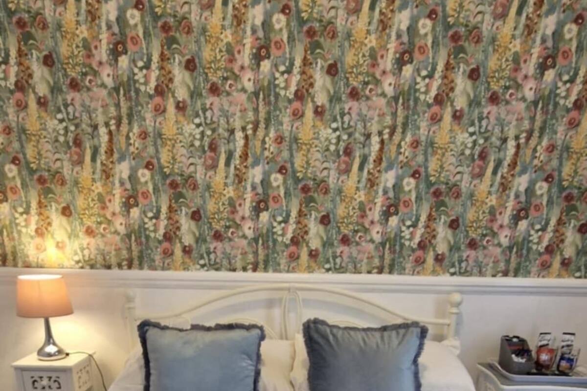 A hotel room with a floral wallpaper, a bed with two pillows, a blue quilt, and two rolled towels.