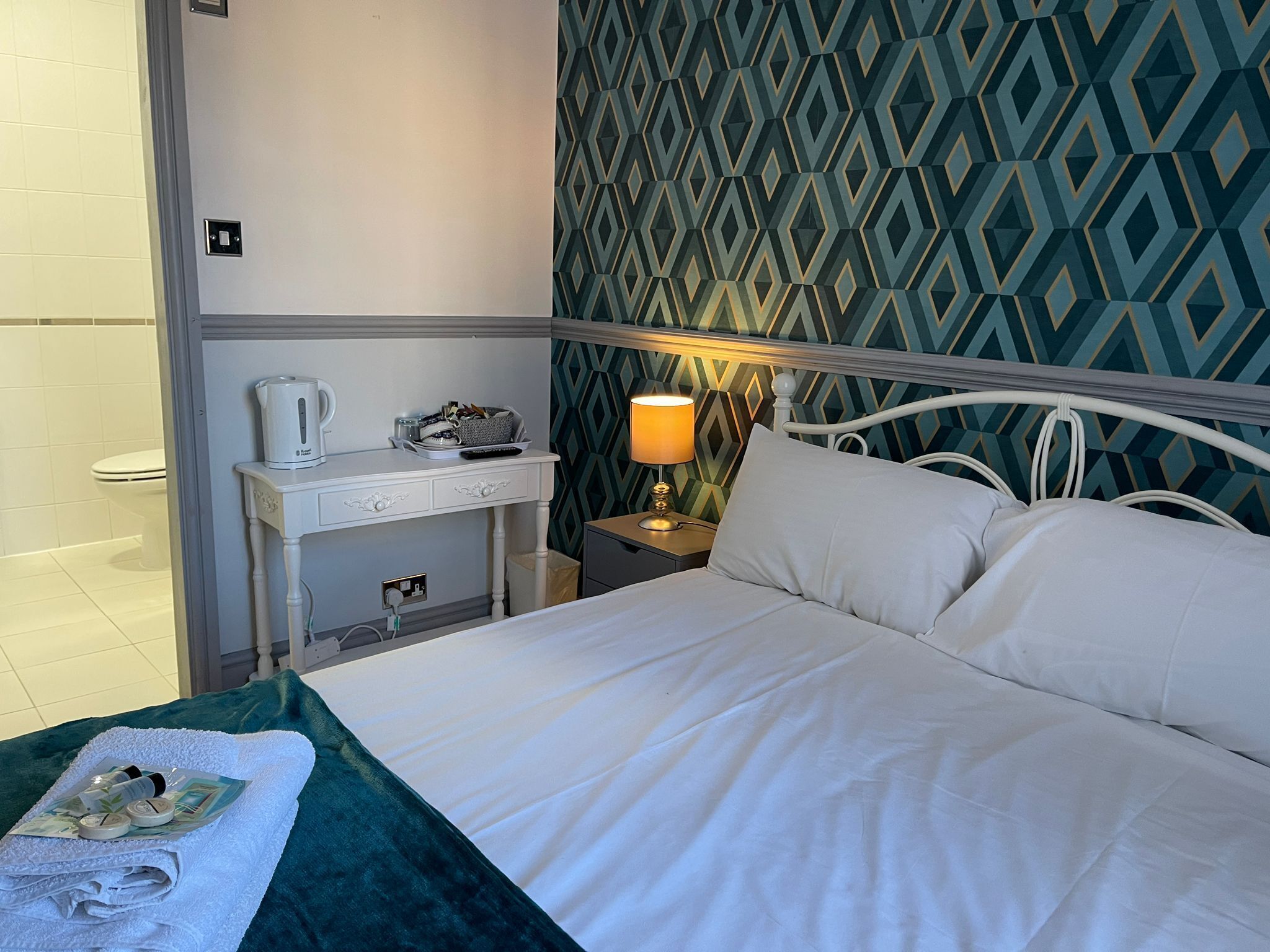 A neatly made bed with white linens, a bedside table with a lamp, and a patterned wall.