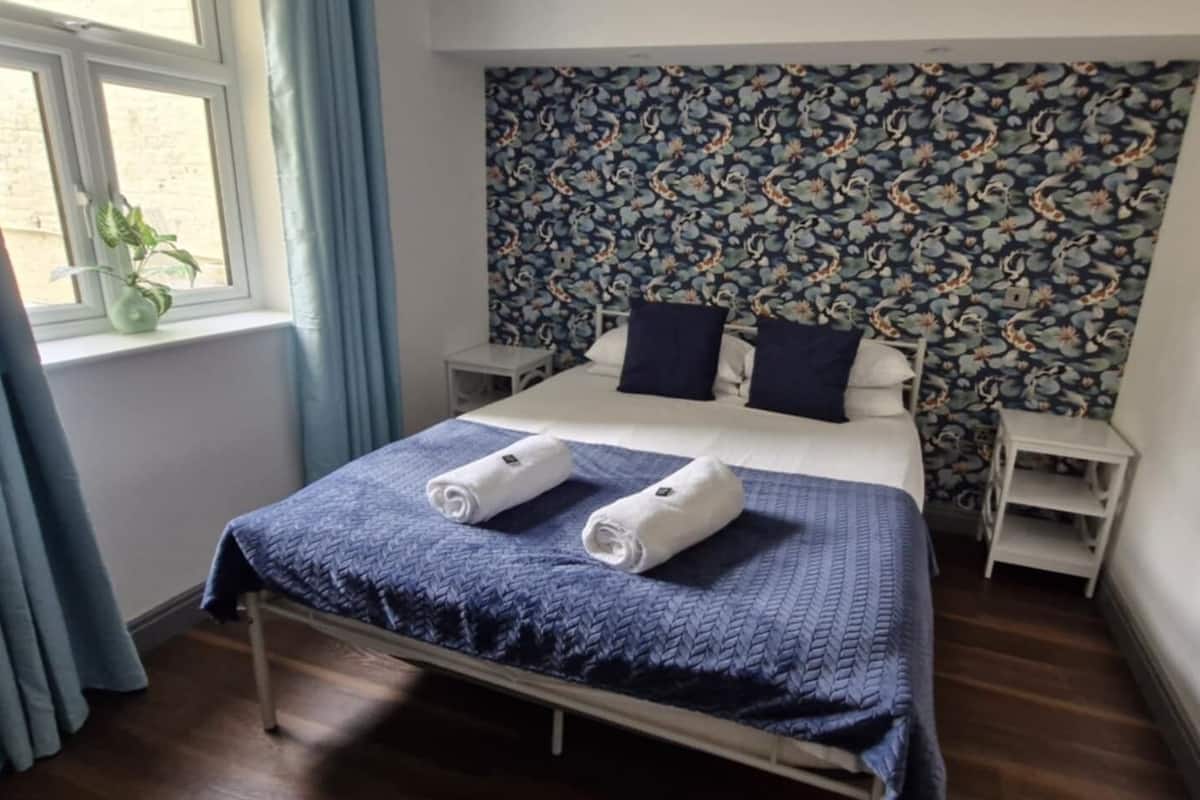 A hotel room with a bed, two pillows, a nightstand, a window with curtains, a floral wallpaper, and a plant.