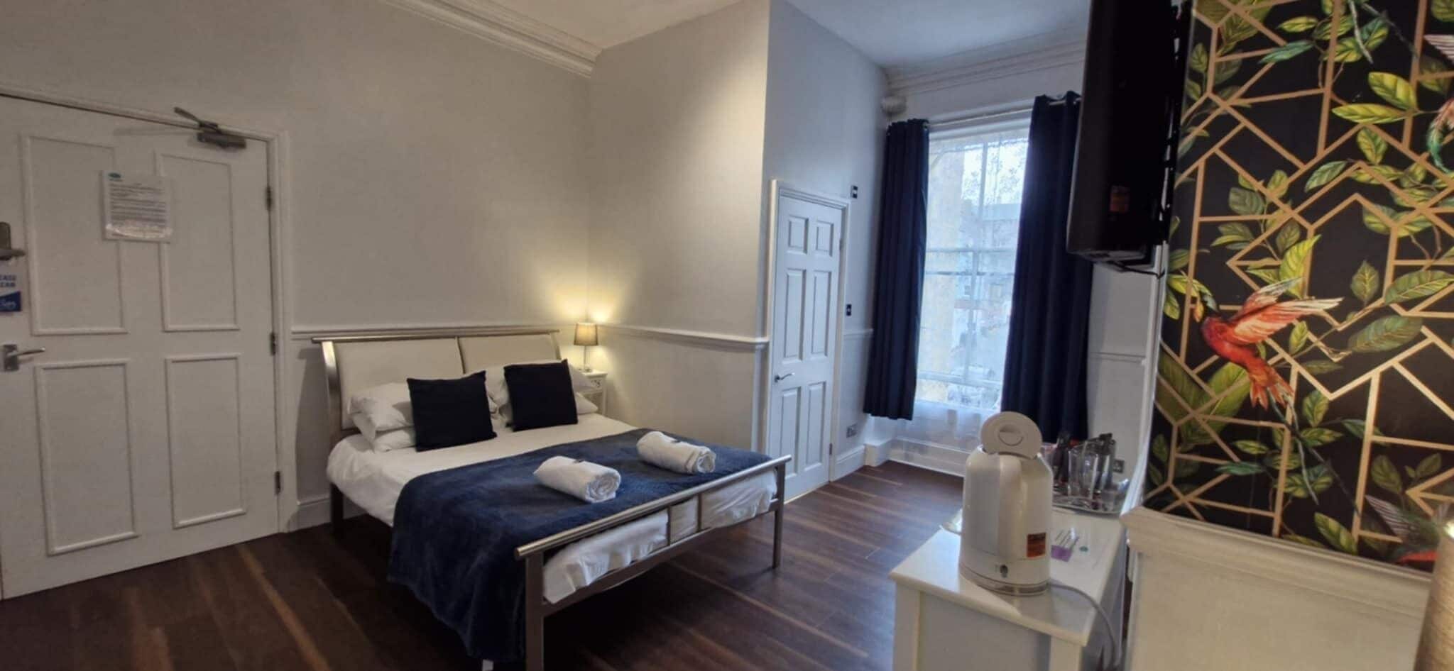 A hotel room with a bed, bedside tables, a desk with a chair, a window with curtains, and a wall with a geometric pattern.